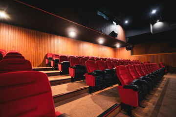 Wide-angle low-key view of empty modern cinema auditorium with rows of red upholstered seats, warm wooden wall panels, soft ceiling lights and dramatic perspective, no people, movie theater atmosphere