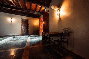 Low-key interior of historic house in Asturias, Salas, featuring rustic wooden ceiling beams, stone tile floor, vintage wall lamps, wooden doors, table and chair, warm light, cozy traditional mood