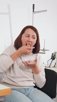 white seamstress eating snack in studio, woman pauses at sewing workstation holding plastic container, picks pieces and tastes food then returns focus to fabric and machine, wearing casual sweater