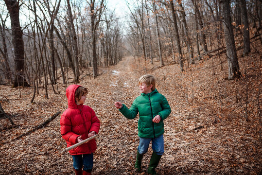 Two young boys wearing jeans, wellies and puffer jackets talking and walking through an autumn forest, USA
