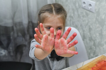 A little girl shows her hands smeared with vegetables in the kitchen. The baby's hands are red from beetroot. The child cooks in the kitchen.