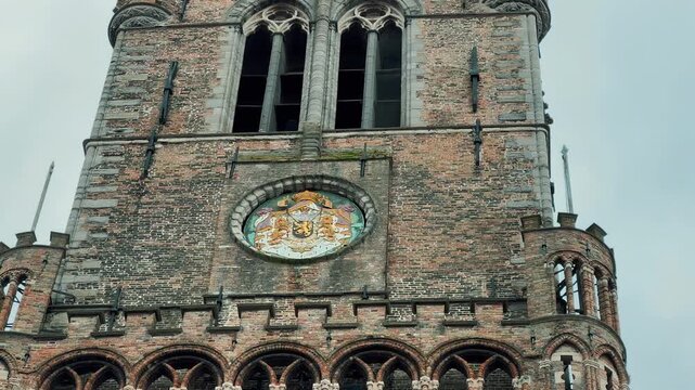 Tilt up view of the medieval Belfry tower in Bruges, Belgium. This historic brick landmark features Gothic architecture, waving flags, and a clock face against a cloudy sky.

