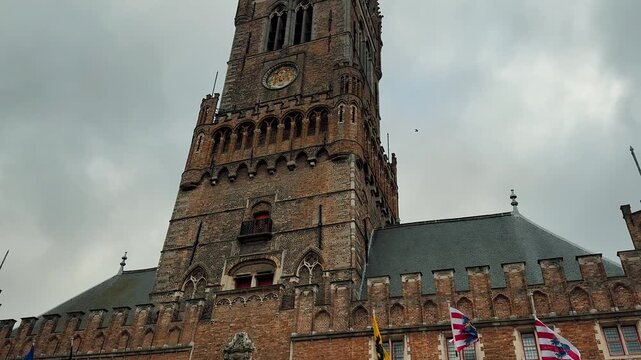 Tilt up view of the medieval Belfry tower in Bruges, Belgium. This historic brick landmark features Gothic architecture, waving flags, and a clock face against a cloudy sky.

