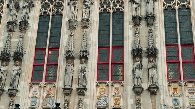 A detailed view of the Provincial Court on the Market Square in Bruges, Belgium. The neo-gothic building features flags waving and intricate stone details on a cloudy day.

