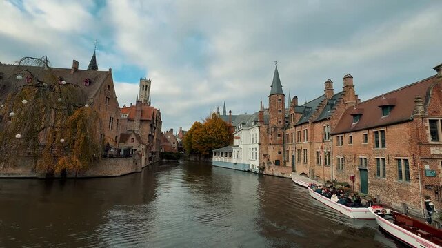 Scenic view of Rozenhoedkaai canal in Bruges, Belgium. Features passing tourist boat, medieval brick architecture, willow tree, and the iconic Belfry tower rising in the background.

