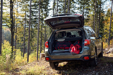 Car is parked on a dirt path in a forest, with many tall trees in the background. The vehicle's trunk is open, revealing bags and other items inside.