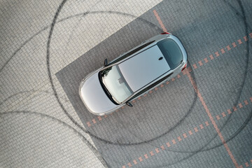 A gray vehicle is parked on a paved lot. Circular black tire tracks are visible on the paving stones, indicating the car performed stunts or circular maneuvers.