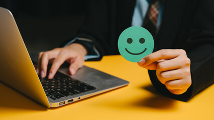 Feedback concept. Person holding green smiley face paper while using laptop on yellow table, symbolizing positive feedback and happiness
