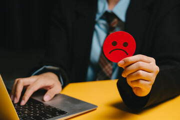 Feedback concept. Unhappy businessperson holding red sad face emoji near laptop on yellow desk showing negative emotion in office setting