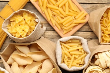 Different types of raw pasta in bags on wooden table, flat lay