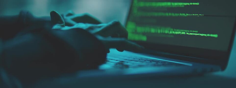 Close-up of a person's hands typing on a laptop keyboard with green coding on the screen in a dark room with blue lighting.