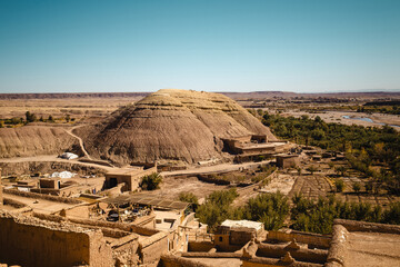 A&iuml;t Ben Haddou, Morocco. Earthen granary and adobe buildings clustered around a rounded hill above a green oasis, desert plains and distant Atlas Mountains under clear blue sky.