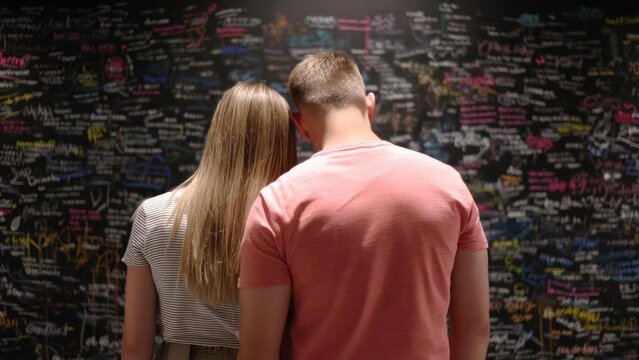 Couple with backs to camera leaning toward a wall covered in colorful chalkboard writing and messages. Concept Backs to camera couple, Chalkboard wall messages, Colorful writing backdrop - Powered by Adobe