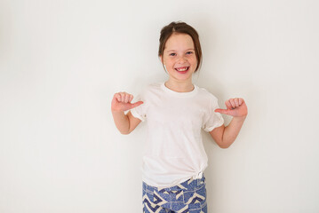 Portrait of a smiling girl standing in front of a white wall pointing her thumbs at herself