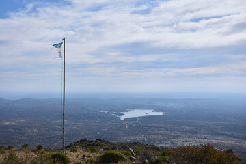 Vig&iacute;a de las sierras: Una bandera argentina antigua flamea en la cima de la monta&ntilde;a con vistas a un lago lejano.