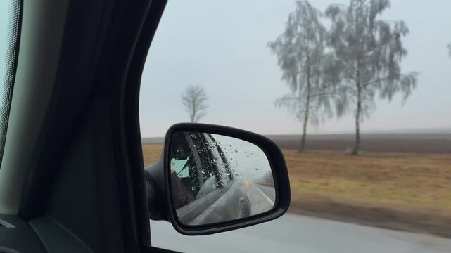 Car side mirror with raindrops, countryside road motion, grey autumn trees, rainy day road trip POV, calm transport b-roll, overcast weather, no people