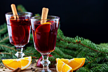 Mulled wine. Festive beverage served in elegant glass, garnished with cinnamon sticks and orange slices, placed on wooden table surrounded by Christmas tree branches, creating warm holiday atmosphere