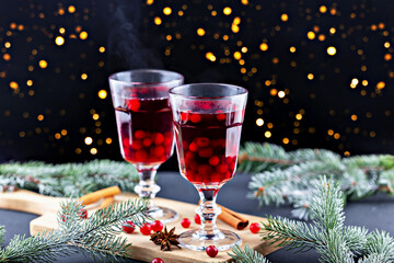 Mulled wine. Two elegant glasses filled with steaming holiday beverages, garnished with cranberries, placed on wooden board surrounded by fresh pine branches, creating warm festive atmosphere