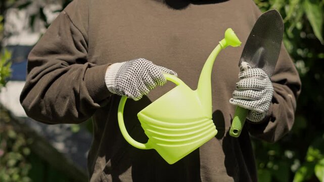 Person holding a small garden trowel in one hand and a watering can in the other, preparing for outdoor gardening and plant care.