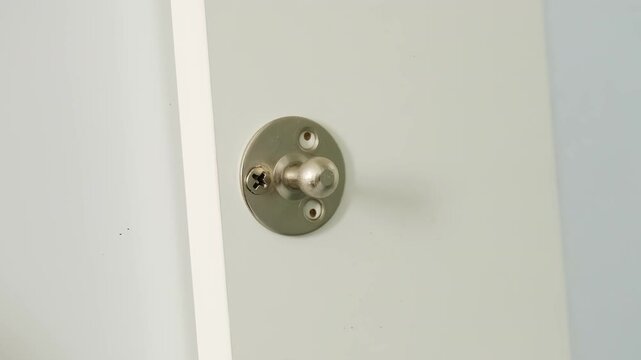 Worker finishes attaching small metal object to surface. Furniture assembler twists metal door handle with index finger and thumb working in well-lit room