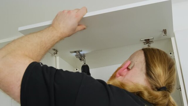 Man tightens screws inspecting furniture and hinges. Craftsman holds onto small wooden cabinet door and checks work done analyzing reliability of fasteners