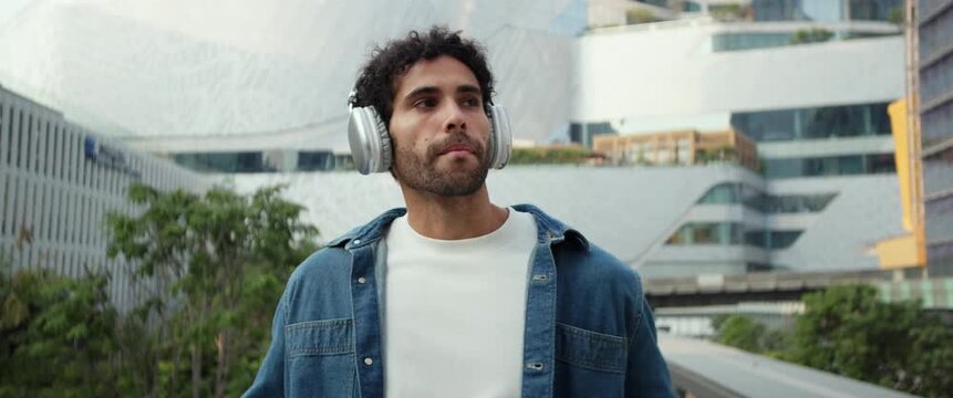 Portrait of bearded Hispanic man puts on earphones to listen to music in city anamorphic lens. Latin guy in denim jacket smiles as hears favorite song in megapolis district