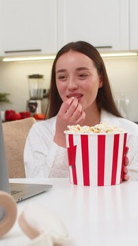 Young woman at home watches comedy trailer on laptop eats popcorn laughs enjoys break without stress. Caucasian girl on sofa sits at table smiles looks at screen fun mood casual evening online video