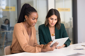 Two engaged multiethnic female colleagues using tablet at workplace, reviewing work application. African professional giving help with job task, feedback, consultation to Latin colleague