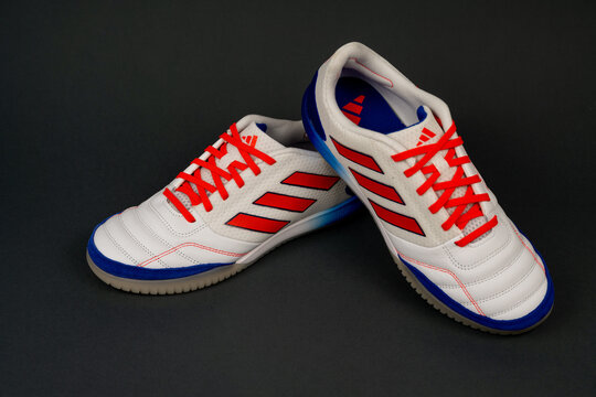 Nis, Serbia - 26 September 2025. A pair of white adidas indoor soccer shoes with red laces and blue accents on a dark background. Modern sports shoes for futsal and training.