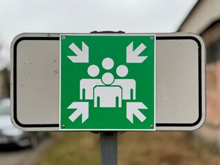 Green emergency assembly point sign with white human figures and directional arrows, indicating a designated meeting area for evacuation and safety procedures.