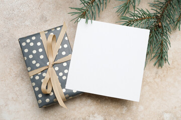 Christmas square greeting card mockup with blank space beside a beautifully wrapped gift, adorned with a ribbon and surrounded by evergreen branches for festive design inspiration