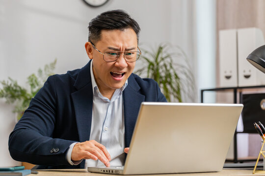 Irritated tired Asian businessman while working on laptop, unexpected online website problem, computer virus data loss by hacking. Freelancer man feeling mad about broken notebook at office workplace
