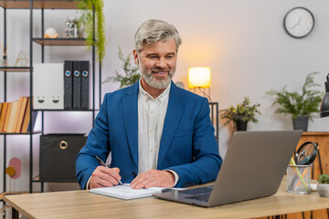 Middle-aged businessman at home office hosts webinar using earphones through laptop presenting topic. Freelancer guy at table answers audience questions sharing expertise guiding learners to success.