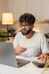 Arabian man at home uses laptop and calculator balances totals enters sums marks paid tracks savings. Egyptian guy sitting at table checks utility bills notes confirms payment plans budget for month.