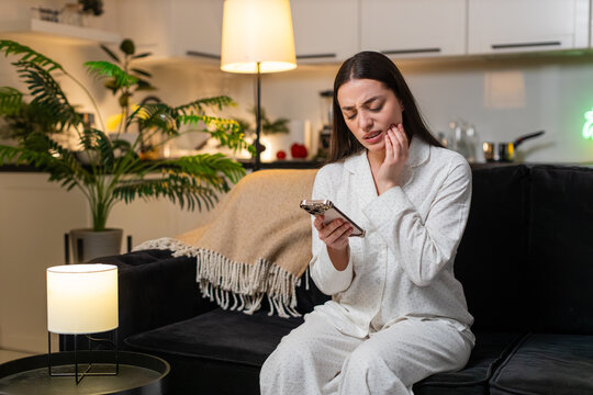 Young woman at night home holds cheek tooth pain uses smartphone to find dentist book visit. Caucasian girl on sofa at evening feels ache frowns types clinic name finds slot taps book time quick help.