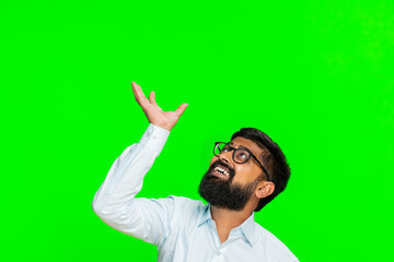 Indian man raises arms looks upward as reaching for floating banner, text or logo, presenting space above. Arabian guy isolated on green chroma key background advertising or promoting virtual content
