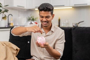Indian man at home drops coins into piggy bank hears ring sound smiles and plans savings for month. Arabian guy on sofa drafts invest plan writes note sets target and records budget for week calmly