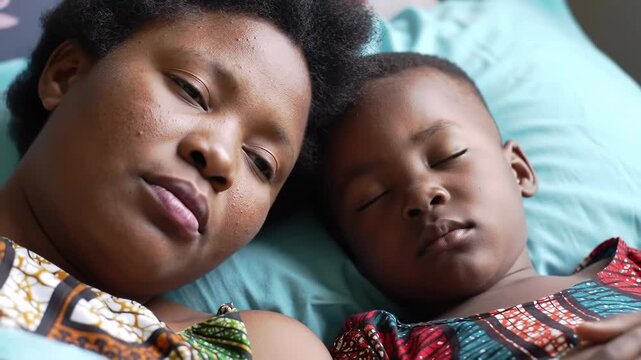 Close up of an African mother and her son napping together on a teal bed wearing Ankara fabric outfits, with soft natural light.