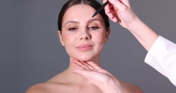Doctor drawing marks on patient's face for plastic surgery operation against grey background, closeup