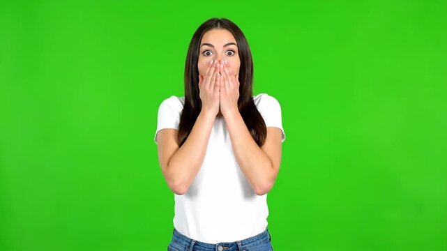 Young woman with a shocked expression covering her mouth with her hands against a green screen background.