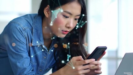 Young Asian woman in denim shirt using a smartphone with abstract geometric overlay. Bright indoor background with a laptop computer on the side. - Powered by Adobe