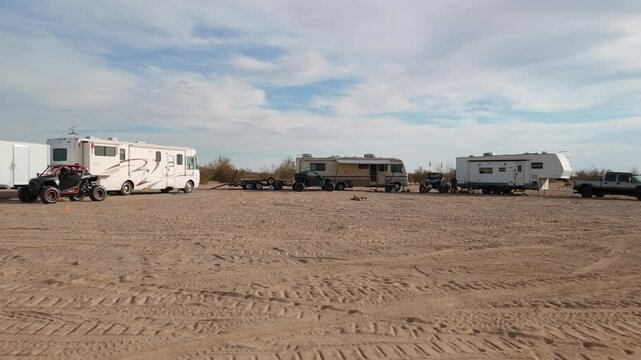 Aerial orbit of a desert camp with motorhomes, trailers, and UTVs parked in a circle.