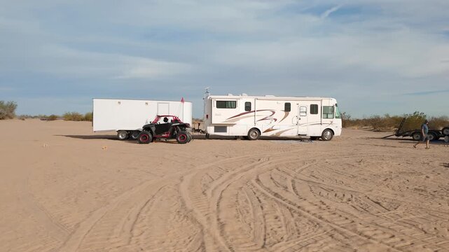 Aerial orbit of a desert camp with motorhomes, trailers, and UTVs parked in a circle.