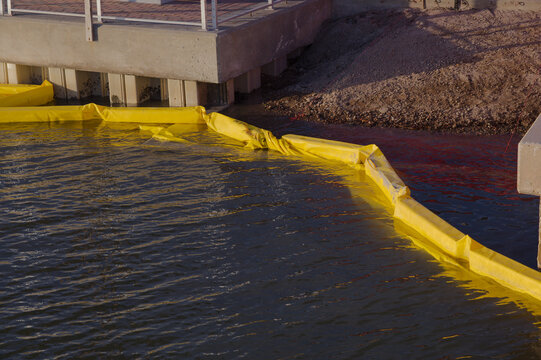 Industrial Waterfront With Yellow Oil Boom water goat Barriers Along Calm Water Near Concrete Pier Structure. Floats along the water beside a concrete pier, illustrating spill response equipment at an
