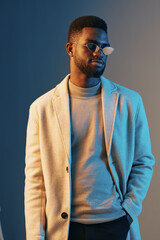 Obraz premium Stylish African American man in a trendy beige coat and sunglasses, expressing confidence against a blue and gray gradient background