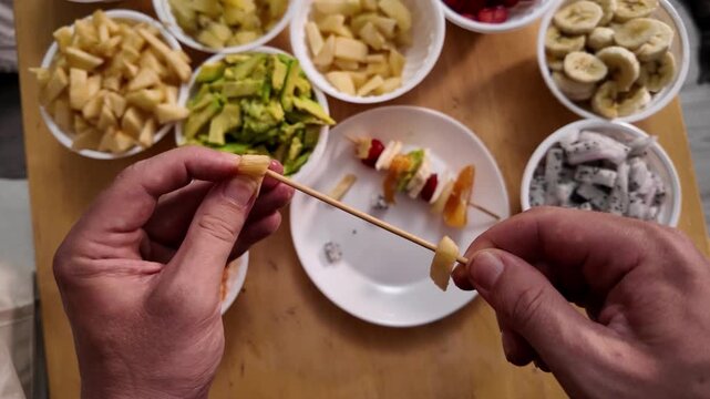 Close-up of men's hands stringing pieces of fresh fruit on a wooden skewer to make fruit kebabs