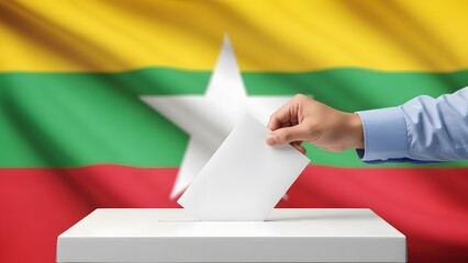 A person's hand casts a ballot into a white election box with the national flag of Myanmar in the background, symbolizing democratic process.