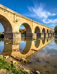 Fototapeta premium A classical stone bridge spans a calm river, reflecting the arches and sky. Clear blue sky with streaky clouds adds to the serene scene