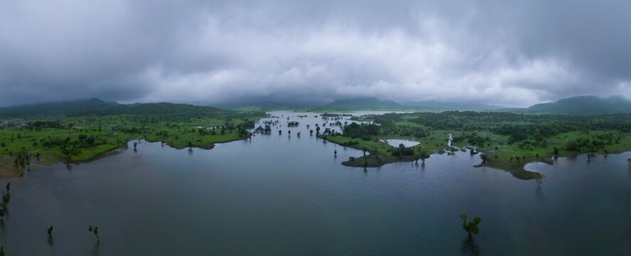 A panoramic monsoon lake view from Bhandardara, Maharashtra, surrounded by lush green hills and misty clouds. Calm water, dramatic sky, and peaceful nature atmosphere.