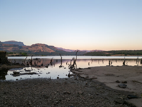 Wide panoramic view of Ghatghar Lake in Bhandardara, showing dry shoreline, calm water, mountain silhouettes and tree stumps at sunset. A dramatic natural landscape scene in Maharashtra.